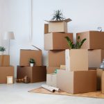 Minimal background image of cardboard boxes stacked in empty room with plants and personal belongings inside, moving or relocation concept, copy space